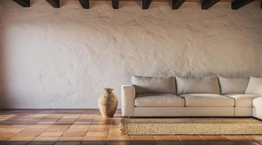 Fotografía editorial de un salón luminoso de estilo mediterráneo español donde conviven una pared encalada de textura rugosa, un sofá de lino natural y una alfombra de fibra vegetal