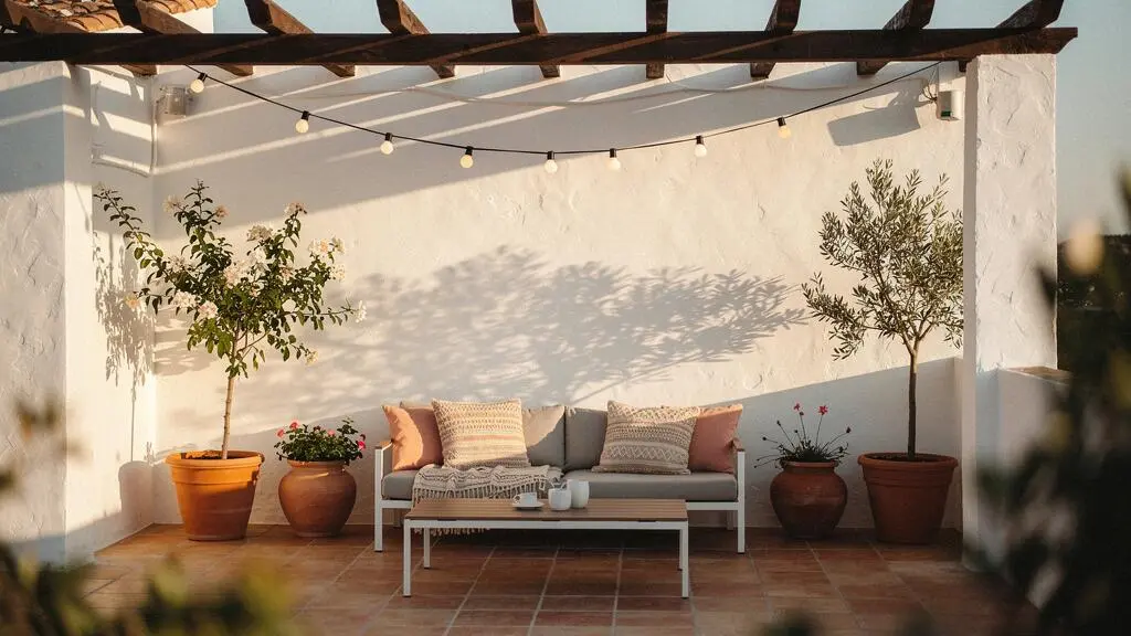 Patio interior español soleado con mobiliario textil, plantas mediterráneas y luz cálida de atardecer, transformado en una acogedora estancia exterior